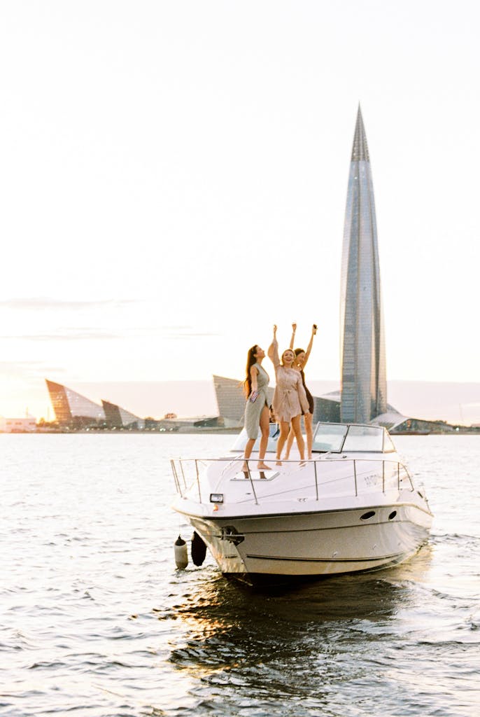 Three women celebrate on a yacht at sunset with a modern tower in the background.
